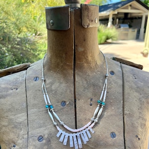 May include: A silver necklace with turquoise beads and a pink, fan-shaped pendant. The necklace is displayed on a wooden mannequin, showcasing its design and craftsmanship. The necklace has a delicate and intricate design.