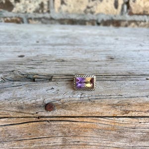 K&ouml;nnte beinhalten: Ein rechteckiger Ring mit einem lila und gelben Edelstein, der in einen silberfarbenen Metallrahmen gefasst ist. Der Ring wird auf einer verwitterten Holzoberfl&auml;che mit einer Backsteinmauer im Hintergrund pr&auml;sentiert.