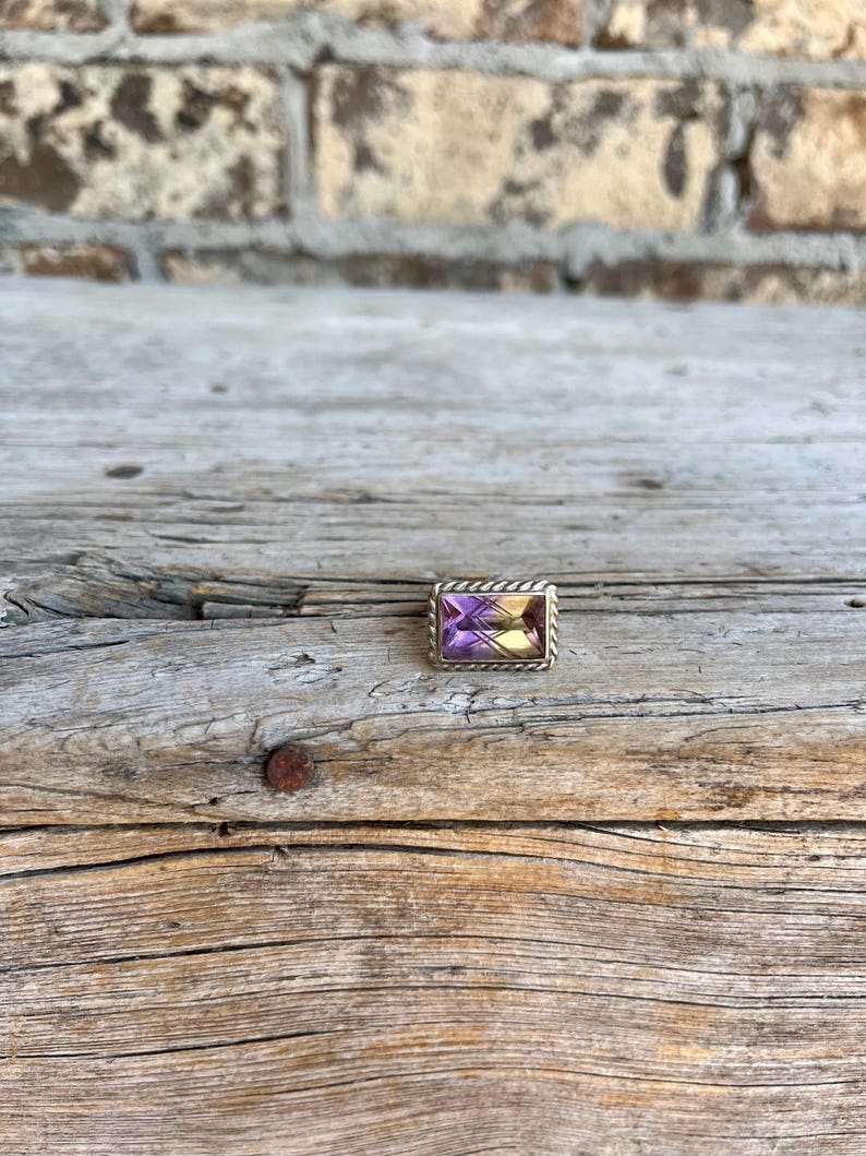 K&ouml;nnte beinhalten: Ein rechteckiger Ring mit einem lila und gelben Edelstein, der in einen silberfarbenen Metallrahmen gefasst ist. Der Ring wird auf einer verwitterten Holzoberfl&auml;che mit einer Backsteinmauer im Hintergrund pr&auml;sentiert.