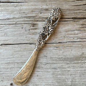 May include: Silver butter knife with a decorative handle featuring a grape and leaf design. The blade is curved and polished, reflecting light. The handle has detailed grape clusters and leaves. The knife is set against a rustic wooden background.