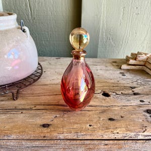 Puede incluir: Un frasco de perfume de vidrio vintage en forma de l&aacute;grima con un tap&oacute;n esf&eacute;rico. La botella es un degradado de rosa y rojo con detalles dorados. El tap&oacute;n es de color dorado transparente. La botella est&aacute; sobre una superficie de madera envejecida.
