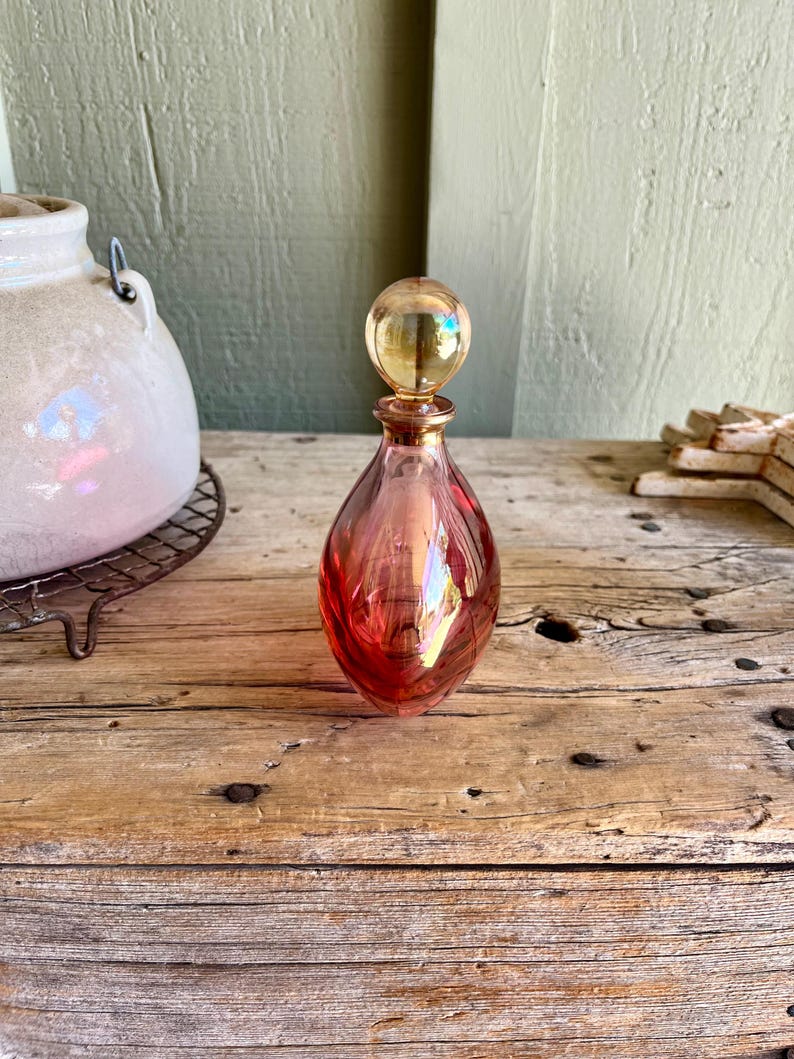 Puede incluir: Un frasco de perfume de vidrio vintage en forma de l&aacute;grima con un tap&oacute;n esf&eacute;rico. La botella es un degradado de rosa y rojo con detalles dorados. El tap&oacute;n es de color dorado transparente. La botella est&aacute; sobre una superficie de madera envejecida.