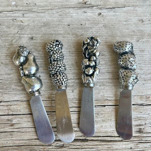 May include: Four silver cheese spreaders with decorative handles. The handles feature designs of a pear, grapes, mushrooms, and strawberries. The blades are rectangular and have text. The spreaders are arranged on a weathered wooden surface.