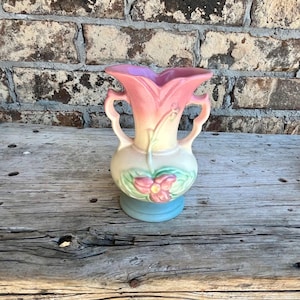 May include: A vintage ceramic vase with a light blue base, a cream-colored body, and a pink and purple flower-shaped top. The vase features two handles and a pink flower design with green leaves. The vase is sitting on a wooden surface.