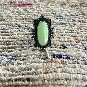 May include: A silver-toned ring with an oval, pale green stone. The ring has an ornate, decorative frame with a beaded edge and scrollwork details. The ring is set against a textured, multi-colored fabric.