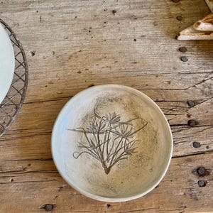 May include: A white ceramic bowl with a simple design of a plant pressed into the surface. The bowl is sitting on a wooden surface.