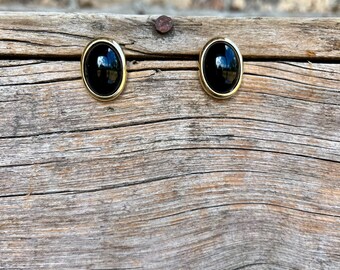 May include: A pair of oval-shaped black earrings with a gold-colored border. The earrings are set against a weathered wooden surface, with a brick wall in the background. The earrings are a classic jewelry accessory.