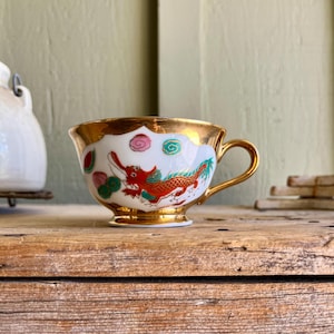 May include: A white teacup with a gold rim and a red dragon design. The cup has pink and green accents and is sitting on a wooden surface.