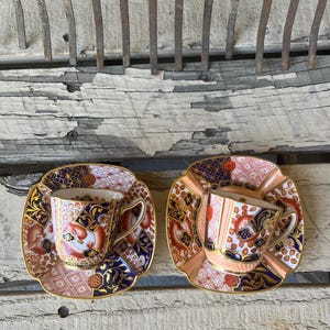 May include: Two vintage teacups and saucers. The cups and saucers feature intricate designs with gold edging, orange, blue, and white patterns. The saucers are square with a scalloped edge. The background is a weathered wooden surface.
