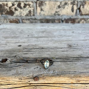 May include: A silver ring with a marquise-cut, light blue gemstone. The ring has an ornate, swirling design on the band. The ring is set against a weathered wooden surface.
