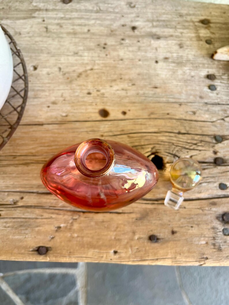 Puede incluir: Una botella de vidrio iridiscente en forma de pera, en tonos rojos y dorados, con una peque&ntilde;a abertura redonda. Un tap&oacute;n de vidrio separado con una base cuadrada transparente est&aacute; cerca. La botella descansa sobre una superficie de madera envejecida.