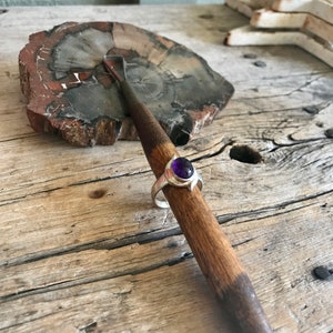 May include: A silver ring with a purple gemstone set in a bezel setting. The ring is resting on a wooden surface.
