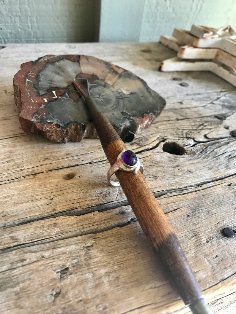 May include: A silver ring with a purple gemstone set in a bezel setting. The ring is resting on a wooden surface.