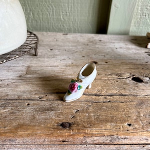 May include: A small white porcelain shoe with a pink rose and green leaves on the toe. The shoe has a gold trim around the edge.