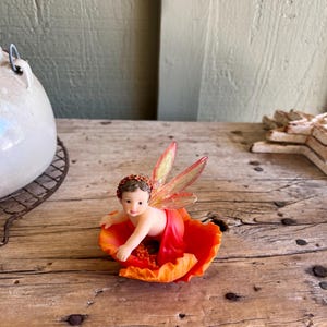 May include: A small fairy figurine with orange wings sits on a red poppy flower. The fairy is wearing a red dress and has a crown of leaves on its head.