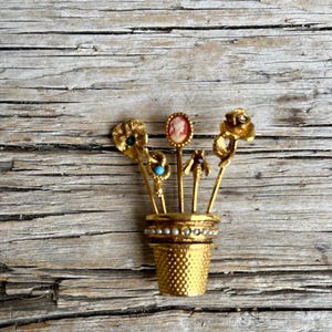 May include: A gold-toned brooch in the shape of a flower pot, holding several decorative pins. The pins feature floral, cameo, and insect designs, with pearl and turquoise accents. The pot has a textured surface.