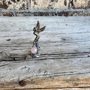 May include: A silver-toned figurine of a winged figure holding a pink sphere. The figure has detailed wings and is adorned with blue accents. The sphere is a soft pink color. The figurine is displayed on a weathered wooden surface.