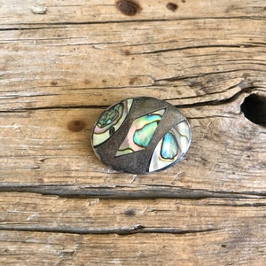 May include: A round silver pin with a geometric design made of abalone shell. The shell has iridescent green, blue, and purple colors.