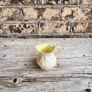 May include: A small cream-colored ceramic creamer pitcher featuring a delicate embossed floral pattern on its body, a pale yellow interior, and a matching pale yellow handle with a curved spout for pouring liquids.