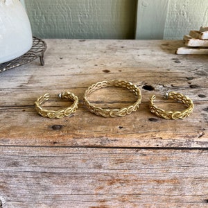 May include: Three gold-toned wire bracelets with a woven design. The bracelets are arranged in a row on a wooden surface.