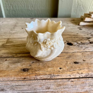 May include: A small, white ceramic vase with a floral design. The vase has a scalloped rim and is decorated with raised roses and leaves.