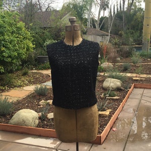 May include: A black sleeveless top with sequins on a dress form. The top is on a stand in front of a garden.