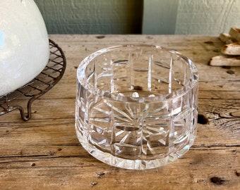 May include: A clear, cut-crystal bowl with a starburst pattern on the base and vertical cuts around the sides. The bowl sits on a weathered wooden surface, with a glimpse of a white ceramic object and a metal trivet in the background.