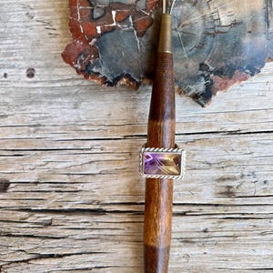 K&ouml;nnte beinhalten: Ein rechteckiger Amethystring mit silbernem, seilartigem Rand wird auf einem Holzwerkzeug pr&auml;sentiert. Der Amethyststein hat Schattierungen von Lila und Gelb. Das Werkzeug hat einen dunkelbraunen Holzgriff und eine silberne Metallspitze.