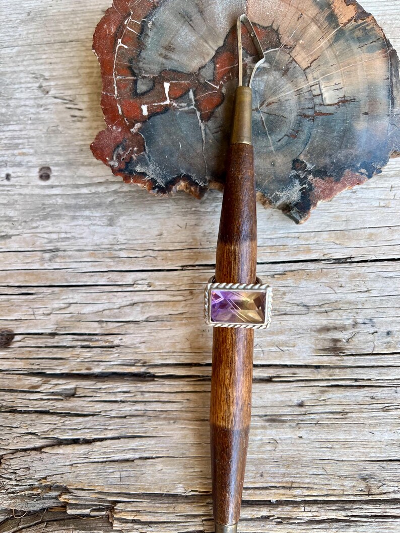 K&ouml;nnte beinhalten: Ein rechteckiger Amethystring mit silbernem, seilartigem Rand wird auf einem Holzwerkzeug pr&auml;sentiert. Der Amethyststein hat Schattierungen von Lila und Gelb. Das Werkzeug hat einen dunkelbraunen Holzgriff und eine silberne Metallspitze.