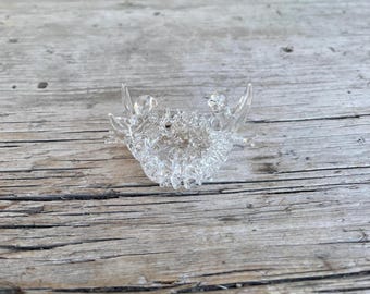 May include: A clear, decorative glass object with a detailed, textured design. The object has a curved shape with two upward-pointing extensions and a central, ornate pattern. The glass is transparent, allowing light to pass through.