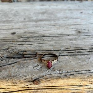 May include: A gold-toned ring with a marquise-cut, deep red gemstone as the centerpiece. The ring is set with smaller, clear stones along the band, creating a sparkling effect. The ring is displayed on a weathered wooden surface.