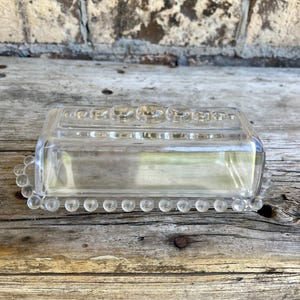 May include: Clear glass butter dish with a rectangular base and a domed lid. The lid and base are adorned with a series of small, round, clear glass beads. The dish is designed to hold a standard stick of butter.