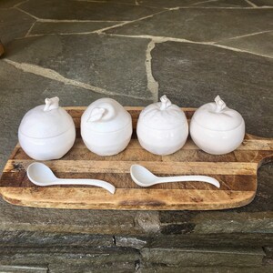 May include: Four white ceramic containers shaped like apples with a small white spoon on a wooden cutting board.