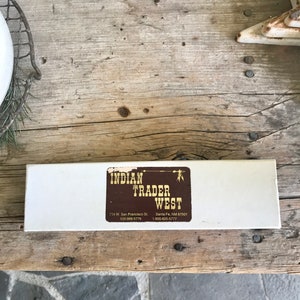 May include: A white box with a brown label that reads "INDIAN TRADER WEST" with contact information for the business. The label also features a stylised drawing of an arrow and a wooden stake.