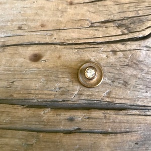 May include: A close-up of a small, round, brass knob with a clear, round crystal in the center. The knob is set against a weathered wooden background.