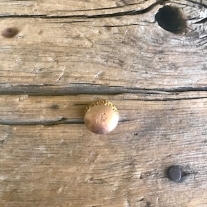 May include: A gold-coloured bottle stopper with a decorative design and the letter "R" engraved on the top.