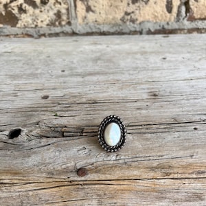 May include: Oval-shaped silver ring with a pearly white center stone. The ring has a beaded border and is set against a rustic wooden surface. The ring is a piece of jewelry.