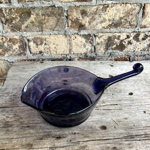 May include: A dark purple glass serving bowl with a handle. The bowl has a curved shape with a pouring spout on one side and a rounded handle with a small ball at the end. The bowl is sitting on a wooden surface.