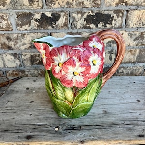 May include: A ceramic pitcher with a floral design. The pitcher features a brown handle and is adorned with red and white flowers with yellow centers, set against green leaves. The pitcher is on a wooden surface.