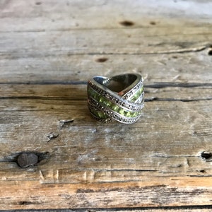 May include: A silver ring with a criss-cross design featuring light green and clear stones.