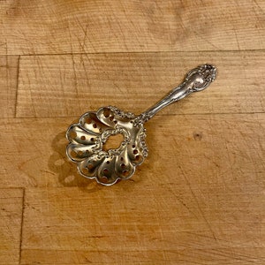 May include: Ornate silver-colored serving spoon with a pierced, fan-shaped bowl and a decorative handle. The spoon features intricate floral and scrollwork designs. A vintage kitchen utensil for serving food.