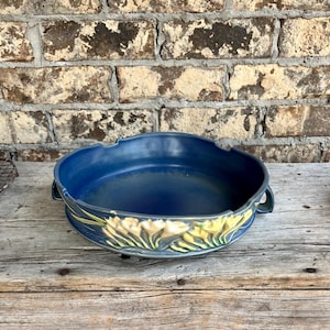 May include: A blue ceramic bowl with a floral design. The bowl has a wide, shallow shape with two handles. The floral design features yellow and green flowers. The bowl is sitting on a wooden surface.