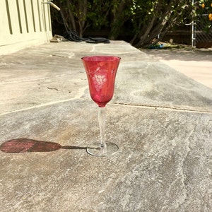 May include: A red glass stemmed wine glass with a grape design etched into the side. The glass is sitting on a gray stone surface.