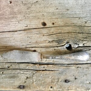 May include: A silver butter knife with an ornate handle sits on a weathered wooden surface. The knife has a dull finish and appears to be vintage.