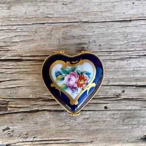 May include: A heart-shaped trinket box with a navy blue exterior and gold trim. The lid features a floral design with pink, red, and blue flowers. The box is set against a weathered wooden surface.