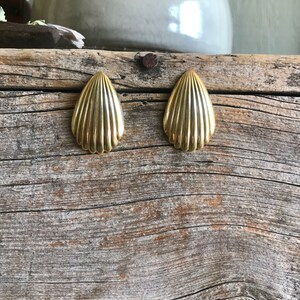 May include: A pair of gold-toned shell-shaped earrings. The earrings are on a wooden surface.