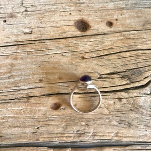 May include: A silver ring with a purple gemstone set in a bezel setting. The ring is on a wooden surface.