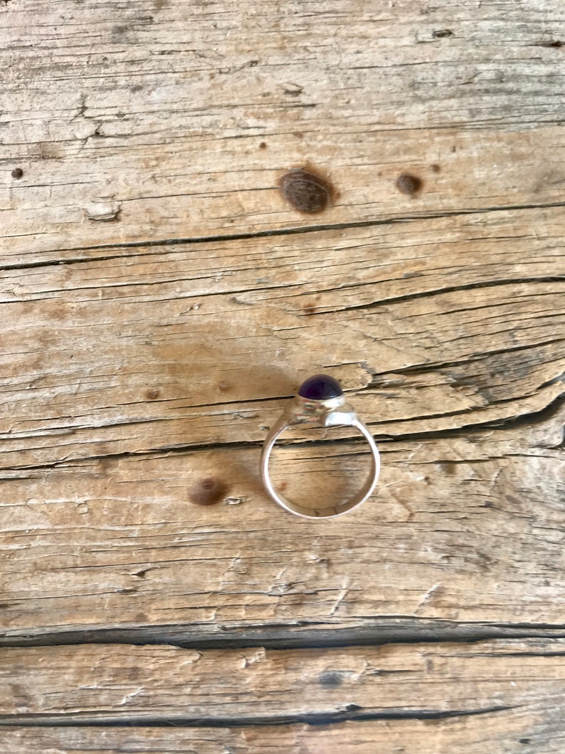 May include: A silver ring with a purple gemstone set in a bezel setting. The ring is on a wooden surface.