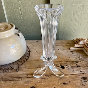 May include: A clear glass vase with a tall, slender body and a flared top. The vase has a three-pronged base with a decorative design.