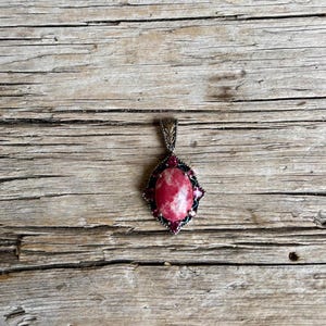 May include: A silver pendant featuring a large, oval, pink and white stone. The stone is encircled by smaller, dark red stones and a decorative silver frame. The pendant is suspended from a silver bail.